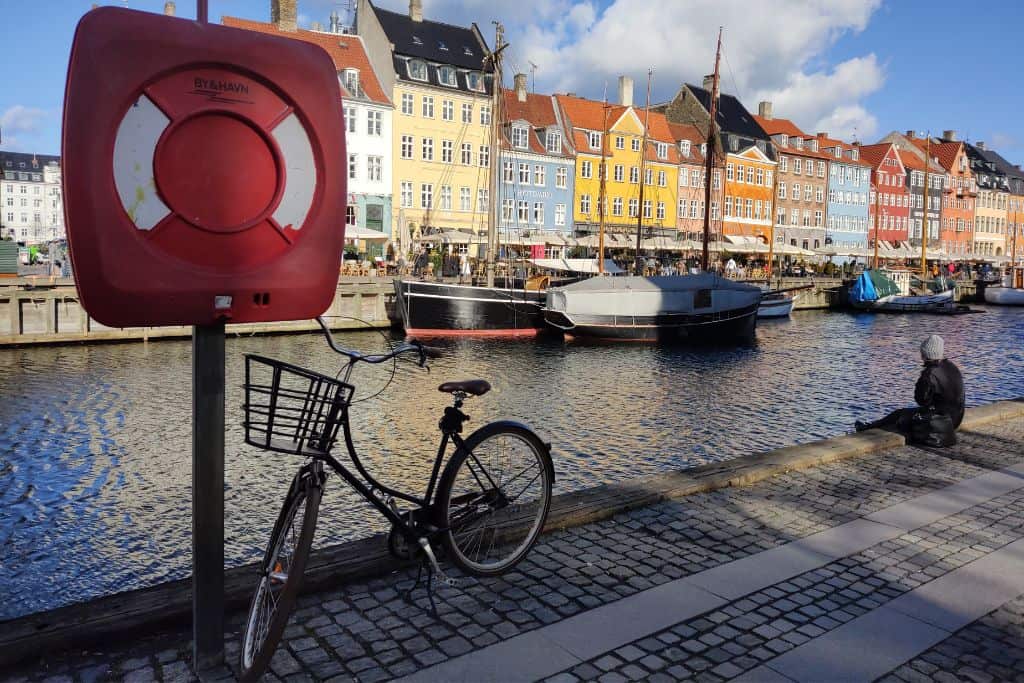 The colourful buildings at Nyhavn, Copenhagen with a bicycle propped up against a lifebuoy and a woman sitting facing the water with her back to the camera.